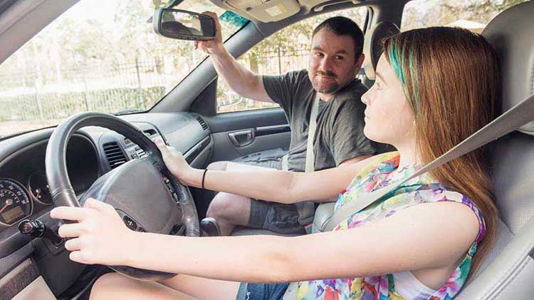 Teen girl in the driver's seat of a car, buckled up with hands on the wheel. Her father is in the passenger seat adjusting the rearview mirror. 