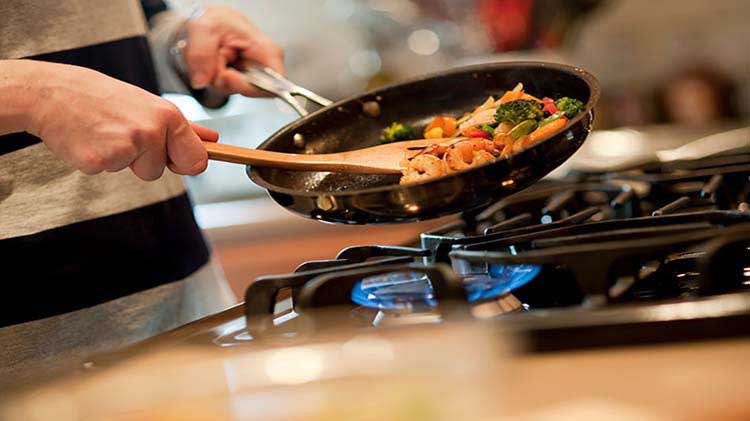 Person holding a pan and using a wooden spoon to saute shrimp and vegetables on a stove with a flame.