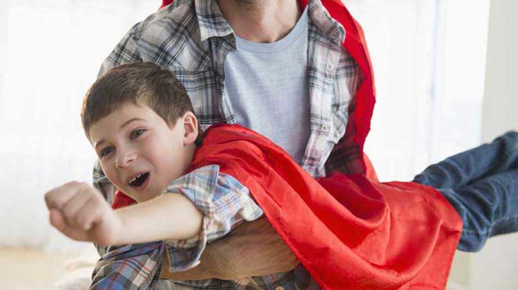 Young boy wearing a cape “flying” in his father’s arms.