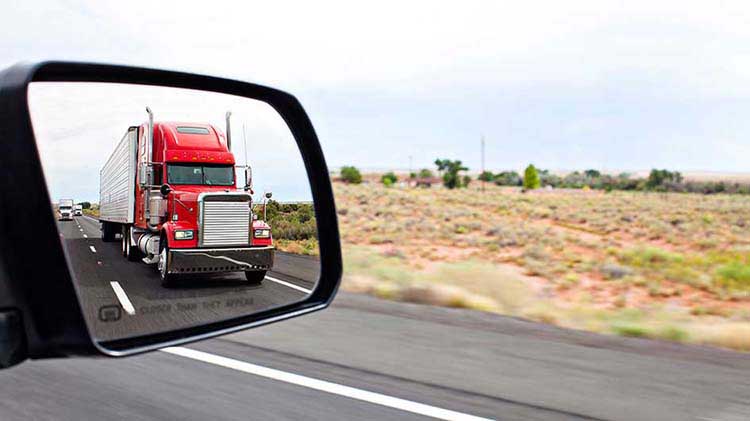 Red semi truck shown in the side mirror of a vehicle.