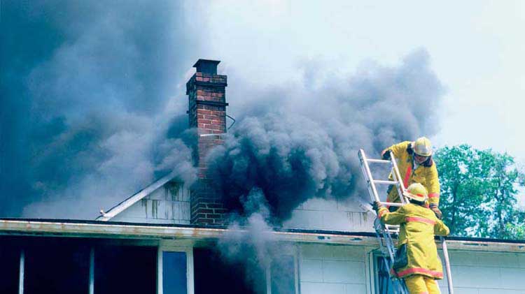 Fire fighters at the top of a ladder preparing to put out a house fire with dark smoke exiting the roof.