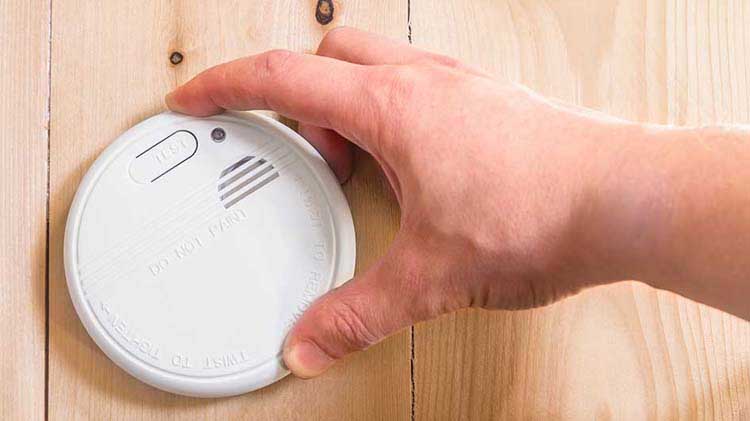 Person's hand holding a smoke detector against a wooden wall.