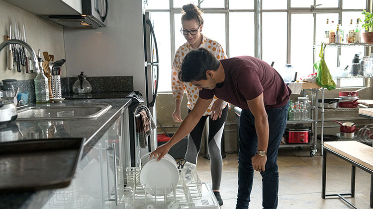 Couple loading dishwasher and discussing appliance maintenance.