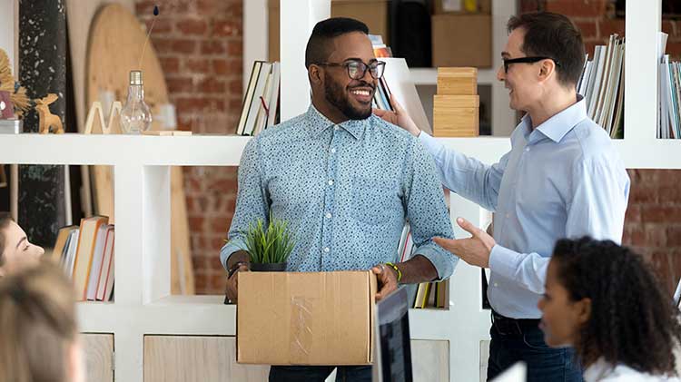 While standing in an office, a man is talking with another man about his 401(k) while carrying a box.