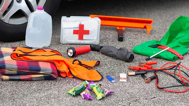 Jug of water, first aid kit, flashlight, umbrella and other emergency kit supplies upacked on the street next to a car tire.