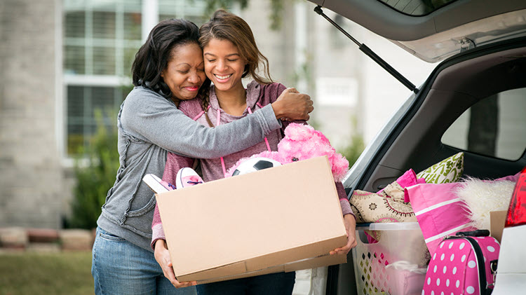 Mom hugging daughter holding box of dorm room supplies.