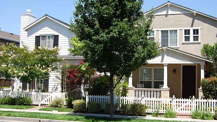 Two homes next to each other in a neighborhood, with white picket fences and trees.