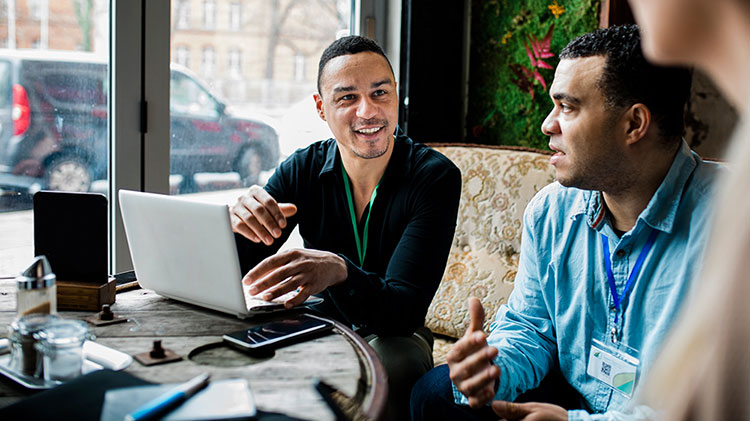 Two men talking and working on a laptop.