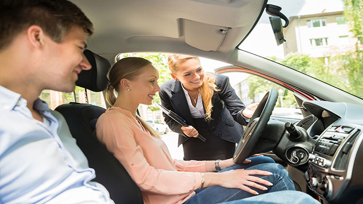 A couple is sitting in a car with the salesperson looking in the window and discussing leasing or buying the vehicle.