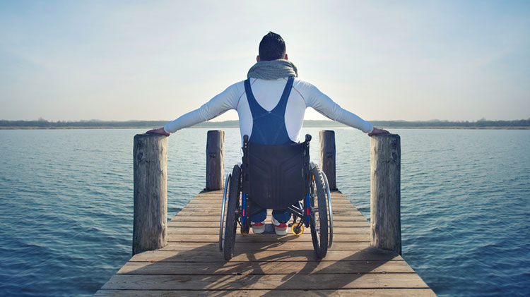 A disabled man sits in his wheelchair on a pier overlooking a lake.