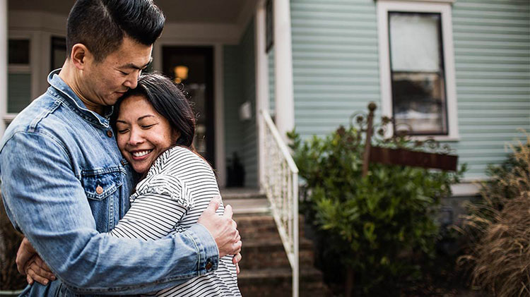 A couple is hugging in front of their home.