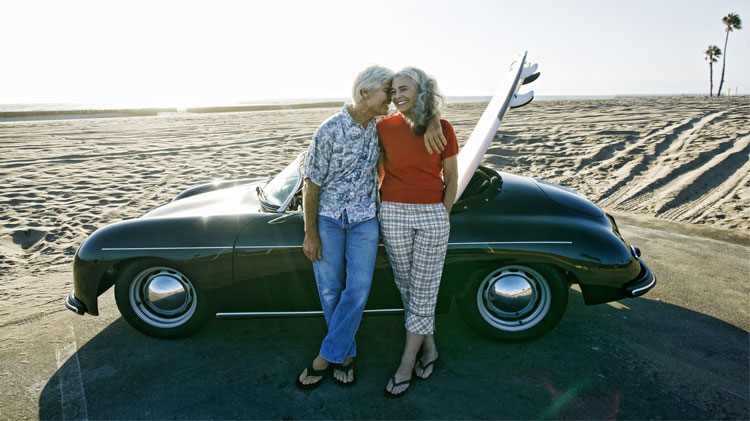 Senior couple leaning on convertible car with a surfboard on the beach.