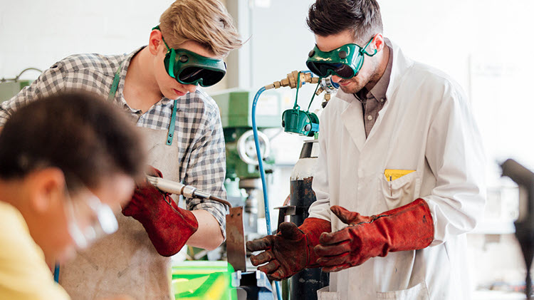 Two young men welding with green safety goggles and red protective gloves.