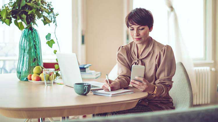 Woman at table, writing on paper while using phone and computer.