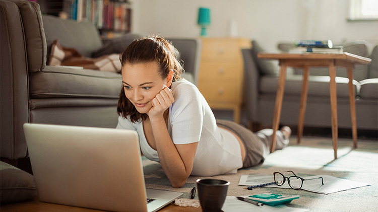 A woman lays on the floor looking at retirement plans on her laptop.